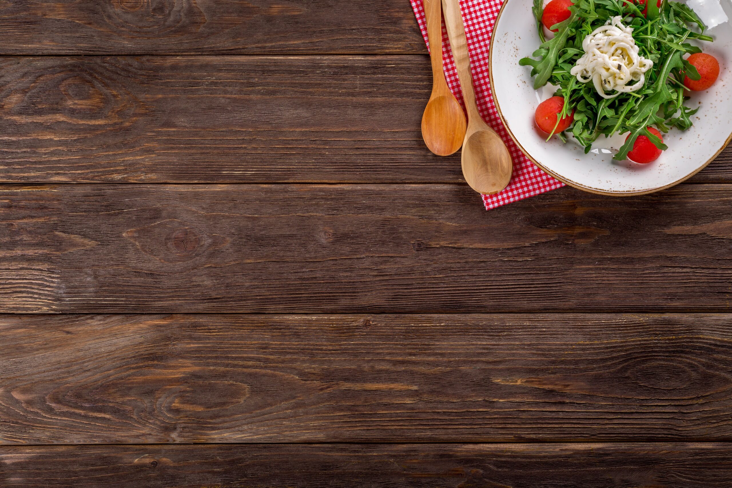 Plate of arugula, tomatoes, and cheese on a wooden table beside wooden spoons and a red checkered napkin.